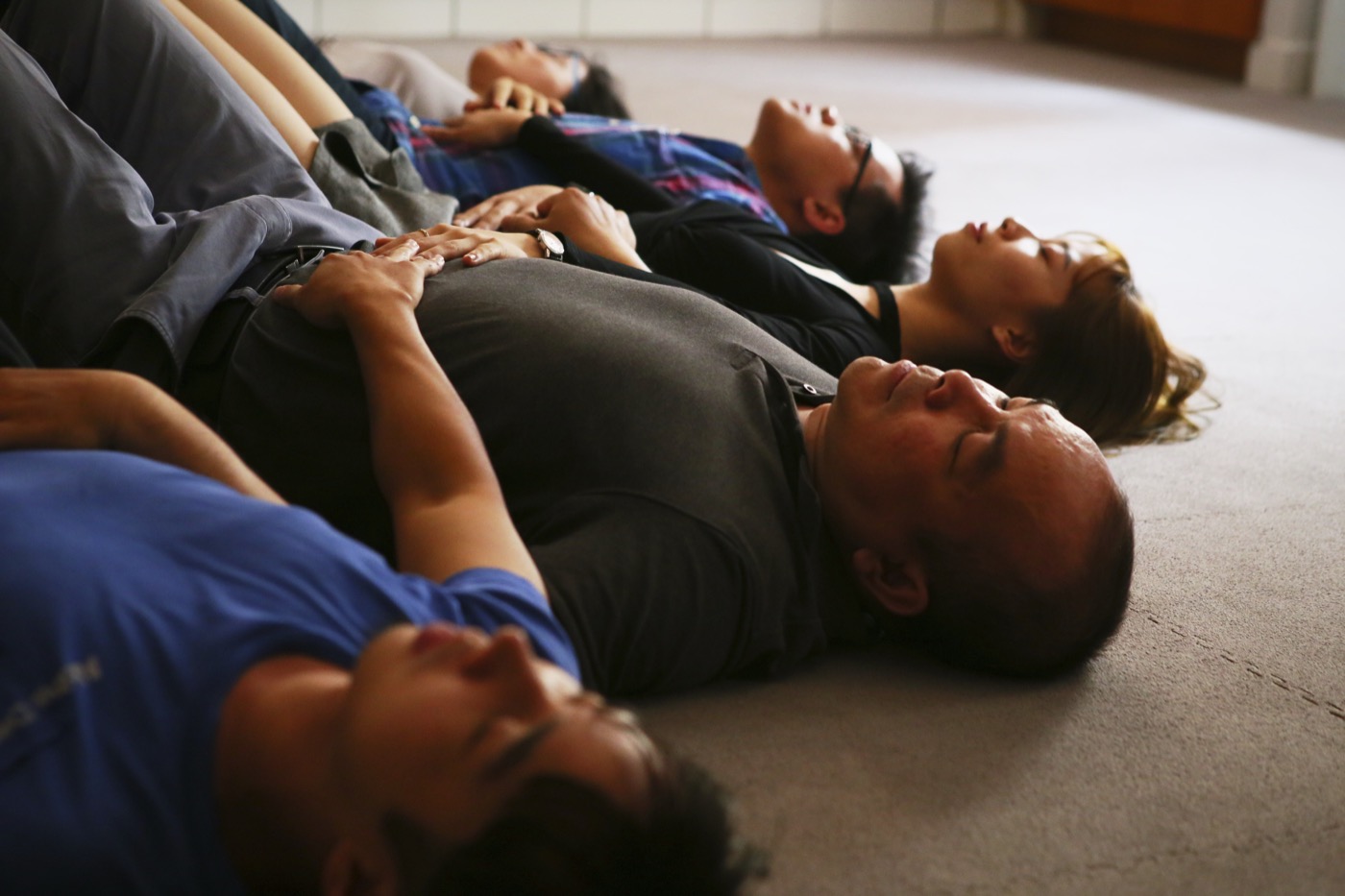 Five adults lying on their backs on a carpeted floor in a row, eyes closed, hands resting on their own chests and ribs, following a breathing exercise