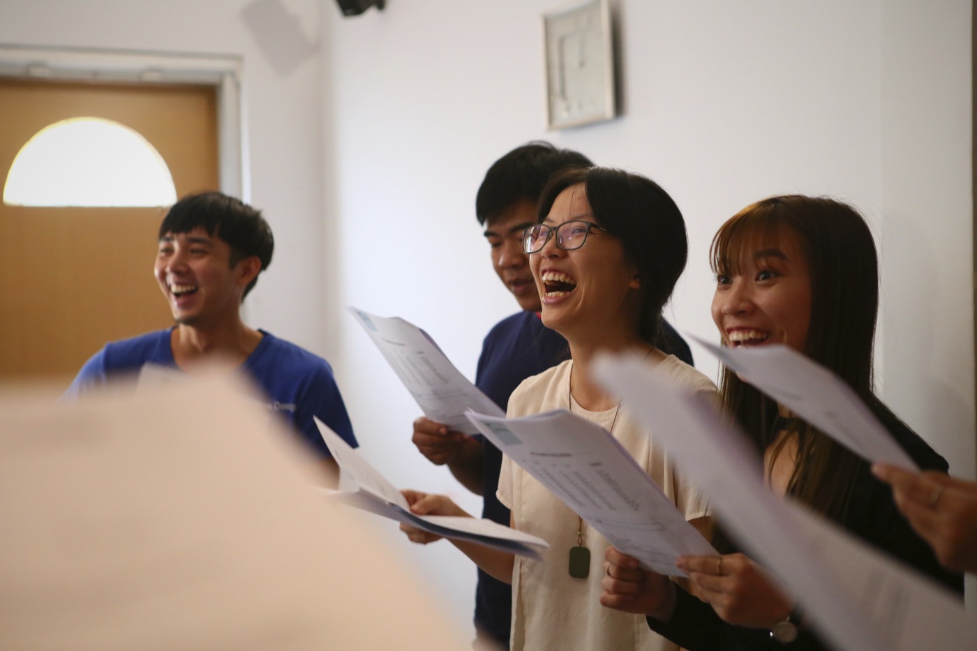 Four people standing in a bright room mid-exercise, worksheets raised, mouths wide open in sound, all of them laughing